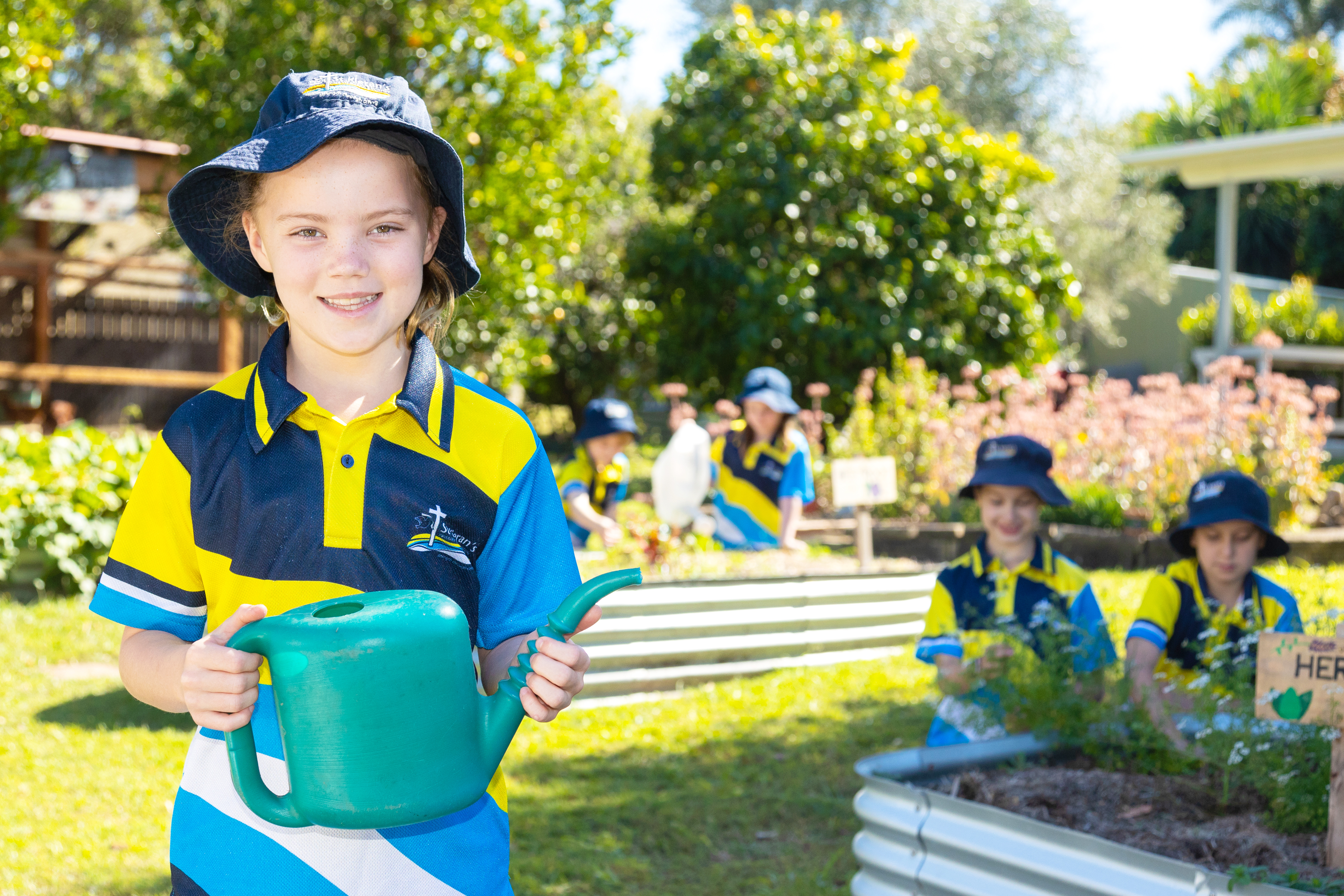 Student in school uniform holding a watering can in a school garden with other students gardening in the background