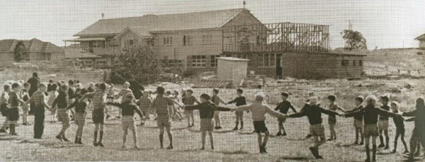 Historic black and white photograph of children holding hands in a circle with a school building under construction in the background