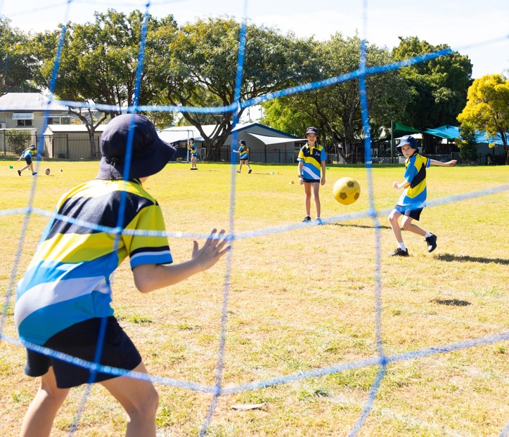 Students in school uniforms playing soccer on a grassy field with one student standing in front of a goal