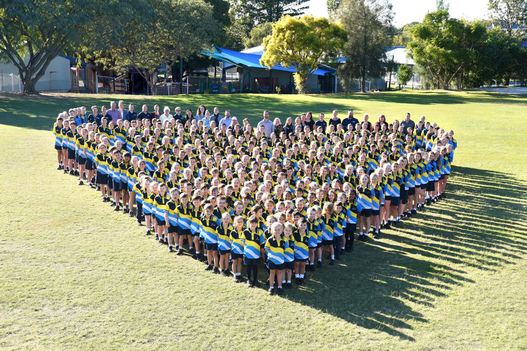 Group of students and staff in school uniforms standing together on a grass field forming a heart shape