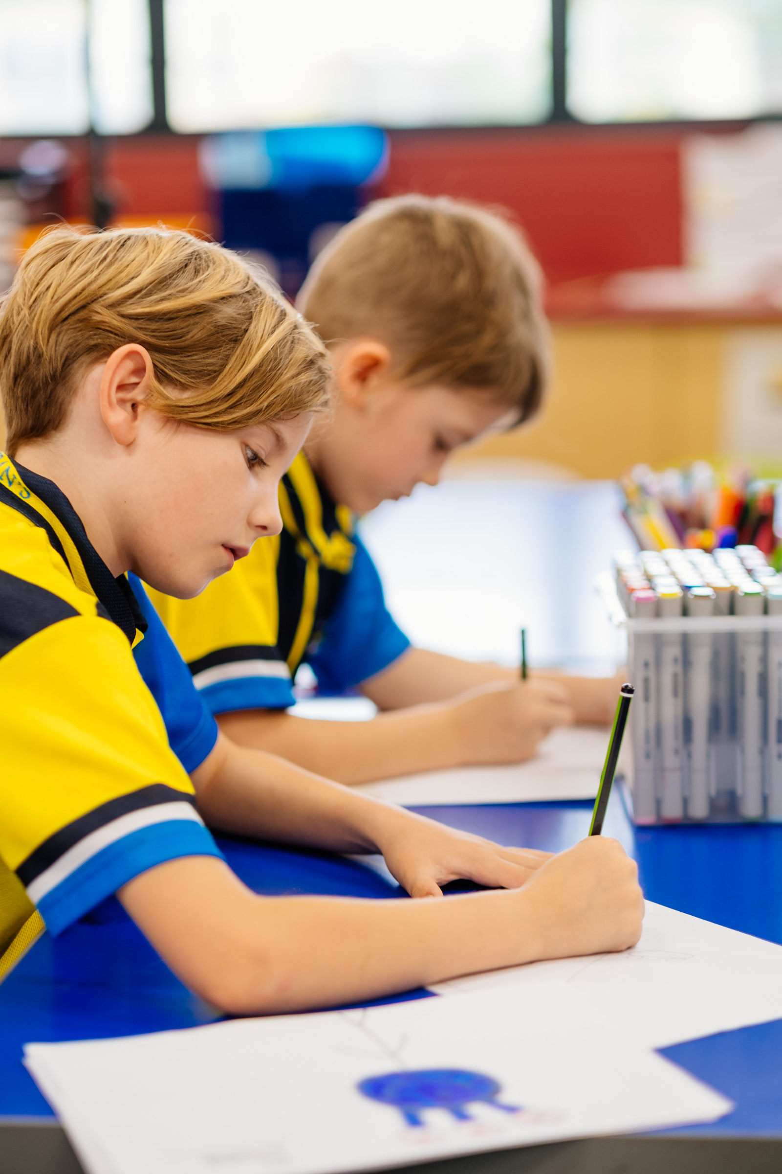Students in school uniforms drawing with pencils at a table in a classroom