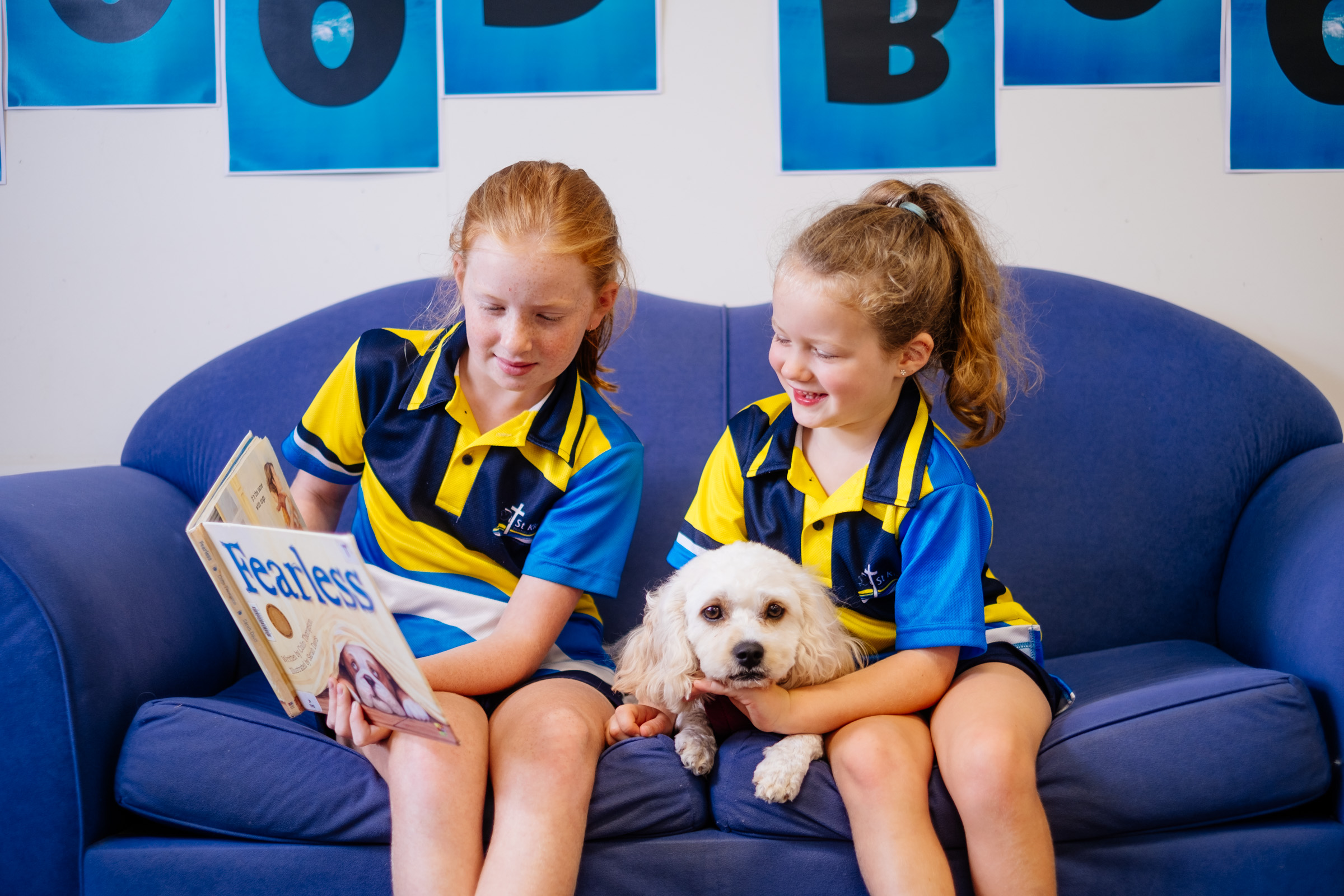 Two students in school uniforms sitting on a couch reading a book with a small dog