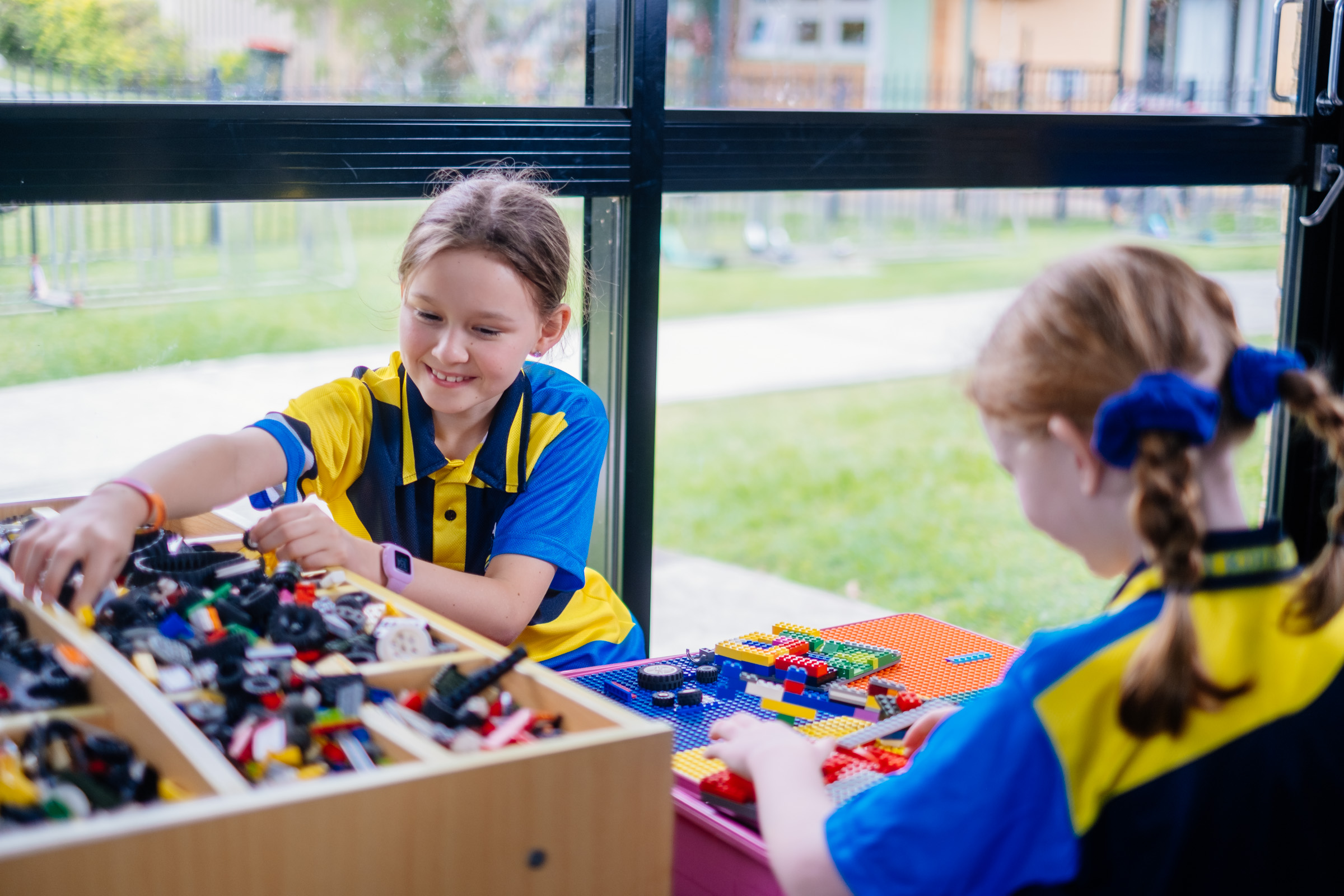 Students in school uniforms building with construction blocks at a table beside a window