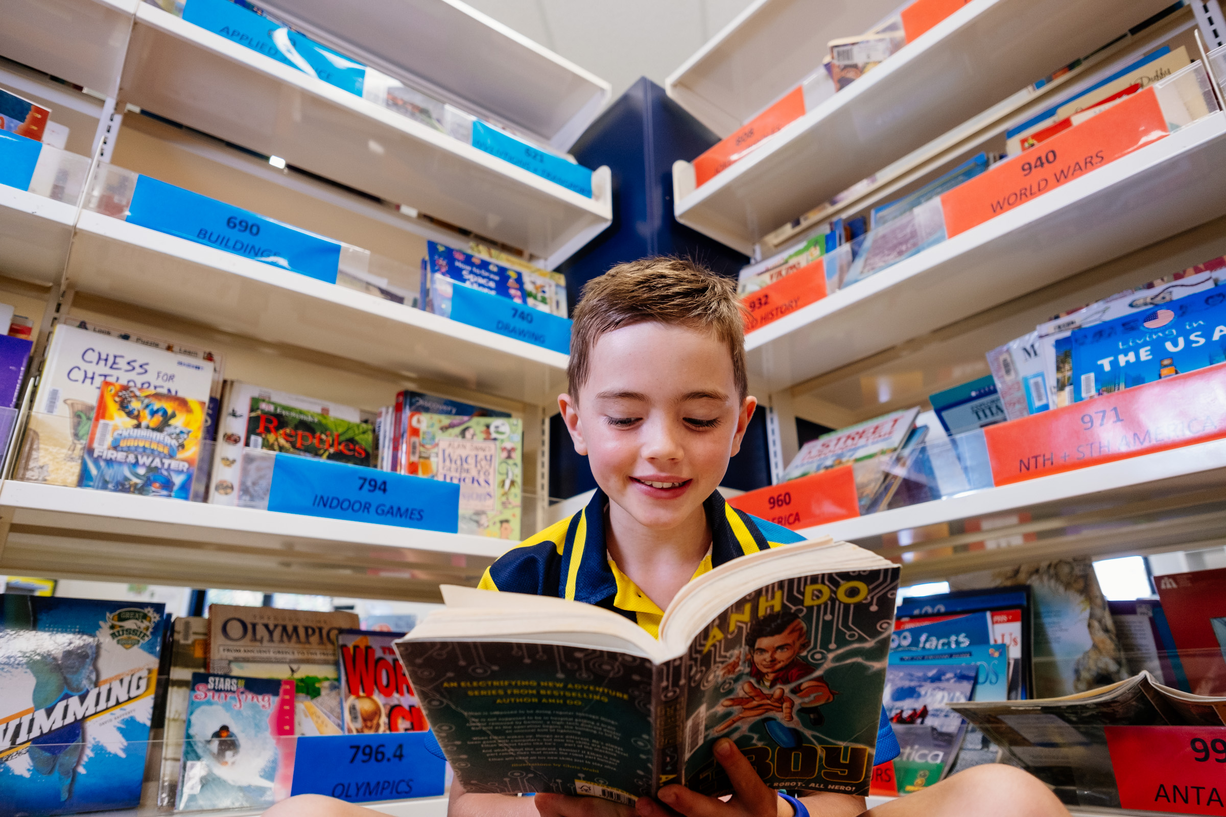 Student in school uniform sitting in a library reading a book surrounded by bookshelves